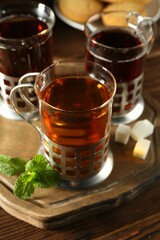 Glasses of hot tea in holders, sugar cubes and mint on wooden table, closeup