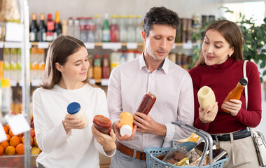 In healthy eating store, positive family of customers chooses sauce and actively argues about purchase. Mom, dad and daughter view variations of gravy, read expiration date of product