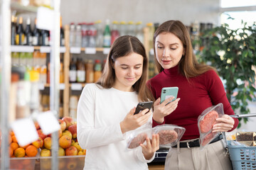 Mother and daughter as a teenager scan packages of meat patties for hamburgers on their phones. Family pays for groceries in a supermarket using a barcode