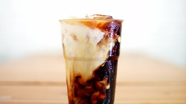 Close-up Move Camera Front view refreshing iced coffee with milk in a clear plastic cup,  swirling blend of dark coffee and creamy milk, Concept coffee art.