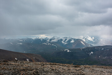 Dramatic top view from high stony pass to alpine valley against large mountain range with snow-capped peaks in gray rainy low clouds. Beautiful big snowy mountain tops in rain under grey cloudy sky.