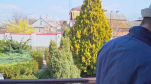 Young man in jacket, hat hangs from balcony of mansion, looks down, runs finger along neck, cold-blooded hand gesture, threatens with violence, revenge, intimidates with mafia shakes head blackmail