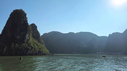 A serene view of Halong Bay in Vietnam, surrounded by towering limestone mountains under a bright sun, with shimmering water reflecting the sunlight.
