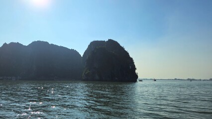 A serene view of Halong Bay in Vietnam, surrounded by towering limestone mountains under a bright sun, with shimmering water reflecting the sunlight.