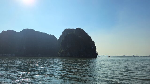 A serene view of Halong Bay in Vietnam, surrounded by towering limestone mountains under a bright sun, with shimmering water reflecting the sunlight.