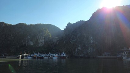 A scenic view of a tourist pier in Ha Long Bay, Vietnam, near Thien Cung Cave, with cruise boats docked by towering limestone mountains under the warm glow of the sun.
