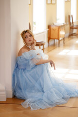 A beautiful young woman in a flowing light blue dress sits tiredly or upset on the floor leaning...