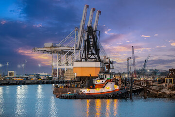 Fototapeta premium Tugboat docked next to cranes in a large port with a cloudscape in the background