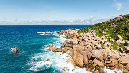 Ocean waters hit rocky coastline under a bright blue sky with lush greenery nearby. La Digue,...