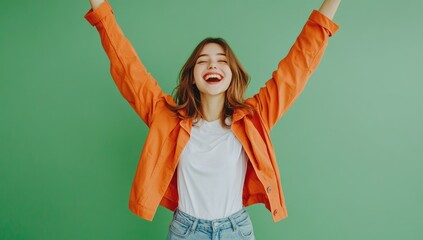 A young woman with brown hair, wearing an orange jacket over a white t-shirt and jeans, joyfully celebrating her success against a green background.