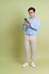 Young Asian man engaging with smartphone while standing against a light green backdrop in a casual yet professional setting