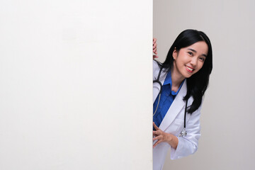 A young female doctor smiling happy while peeking from behind blank wall