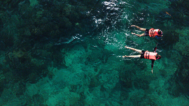 Aerial shot of two snorkelers wearing life jackets floating above a vibrant coral reef in crystal-clear turquoise water. 