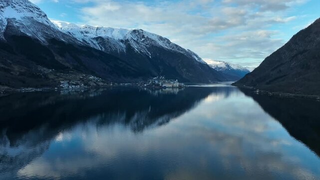 Aerial flying over calm sea in Sorfjorden Norway at dawn. Snow capped mountains, reflections, and industry at Eitrheim