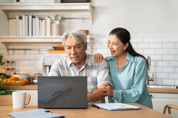 Asian senior man working with laptop at desk by supporting from cheerful wife at home. Couple spending time together.