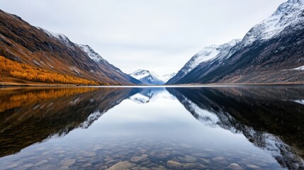 Fototapeta premium A tranquil lake reflects majestic mountains and autumn colors, creating a serene landscape under a cloudy sky.