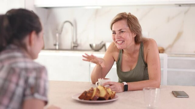 Smiling middle-aged woman talking friendly to her female collaborator while drinking tea in the kitchen