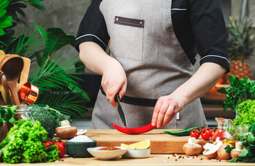 Chef cuts hot red chili pepper. Cozy kitchen with wooden table, kitchenware, vegetables, herbs and ingredients for cooking. Healthy vegan eating, culinary, recipes, food blogging
