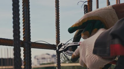 A construction worker is using gloved hands to secure rebar with wire ties at a building site. The clear sky and ongoing construction indicate a busy workday, showcasing the labor involved.