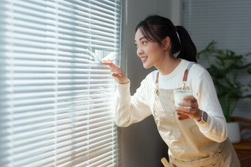 Smiling woman opening venetian blinds and holding glass of milk