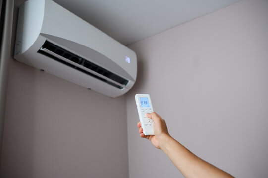 Woman holds an air conditioner remote control with display showing 28 degrees Celsius, sets temperature to heat room, split system hangs on wall. Concept heating with air conditioning