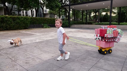 Cheerful young boy pulling small truck loaded with wrapped boxes, presents or Christmas gifts, accompanied by tired dog wearing whimsical bunny ears. Slow motion shot of funny surprises delivery team