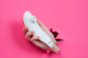 Female hand sticks out white vacuum clitoral vibrator through pink paper background. 