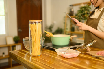 Dry spaghetti in a glass jar, meat, and boiling pot on kitchen counter with woman checking recipe on digital tablet