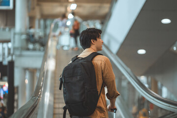 Rear view - Asian tourist man traveller with simple suitcase stand on moving walkway in airport terminal, Tourist journey trip concept.