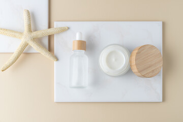 Anti-aging skin care: hyaluronic serum dropper glass bottle and cream jar, dried natural sea star on white marble board over neutral beige background. Top view cosmetic spa card.