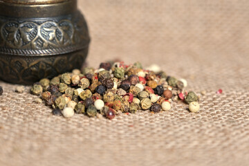 A vintage brass pepper grinder sits on burlap fabric, accompanied by a colorful mix of peppercorns....