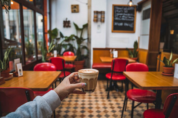 Coffee cup held in cozy cafe with red chairs and greenery in daylight
