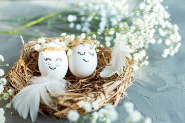 Two chicken eggs with hand drawn happy faces with wreaths in a nest of dry grass with feathers and...