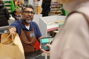 High angle view at male cashier assisting customer paying vis smartphone at supermarket checkout