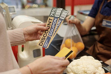 Closeup of unrecognizable female customer handing discount coupons and credit card to cashier at...
