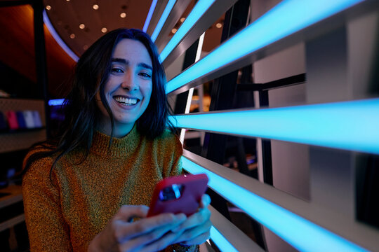 Woman smiling while using phone with futuristic LED lights indoors