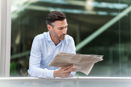Man in business shirt reading a newspaper indoors with a serious expression