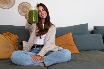 Happy woman showing healthy green juice looking at camera sitting on the couch. Female enjoying...