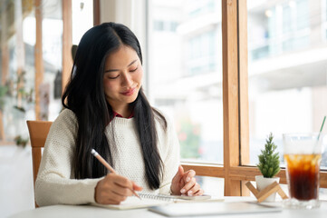 An asian woman is smiling while writing in the notebook, sitting at the table beside window walls.