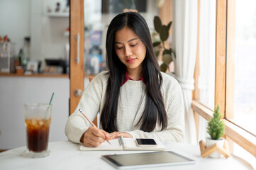 An asian woman is writing with a pencil in notebook with a smartphone and a tablet placing beside.
