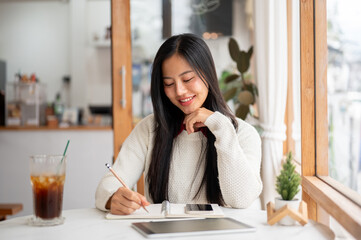 An asian woman is smiling while writing with a pencil on notebook with phone and tablet on the table