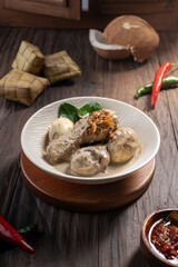 Opor ayam, a rich chicken dish in coconut sauce with eggs, shallots, and ketupat. A spicy and creamy meal for Eid, served on a wooden table with chili and kaffir leaves in the background