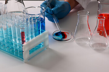 Gloved hand using pipette adding drop of reagent to petri dish with colorful swirl, beakers and...