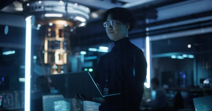 Asian Software Developer Using a Laptop to Monitor the Work of a Quantum Computer, Focusing on Data Flow, Compute and Hardware Stability. Futuristic Dark Setting with Advanced Technological Office