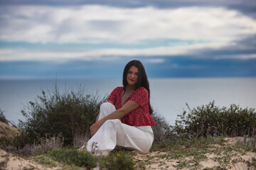 Beautiful young brunette woman is barefoot and sitting on the dunes by the beach. The woman is...