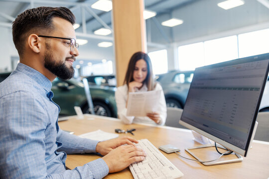 Smiling car seller working with paperwork on computer while lady buyer reading a contract at car showroom.