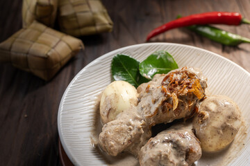 Opor ayam, a rich chicken dish in coconut sauce with eggs, shallots, and ketupat. A spicy and creamy meal for Eid, served on a wooden table with chili and kaffir leaves in the background