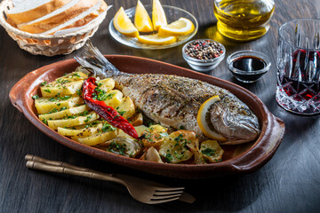 Appetizing baked dorado fish with potato, lemon, red chili pepper and spices in a clay plate, closeup. Healthy food concept, roasted dorado with garnish. Whole grilled fish for eat