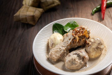 Opor ayam, a rich chicken dish in coconut sauce with eggs, shallots, and ketupat. A spicy and creamy meal for Eid, served on a wooden table with chili and kaffir leaves in the background