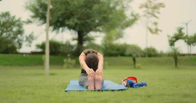 Indian young adult gen z woman lady sitting on yoga mat do stretch warm up toe touch asana in fresh air outdoor garden cute teen female girl lean body learn good self health care habit day time park